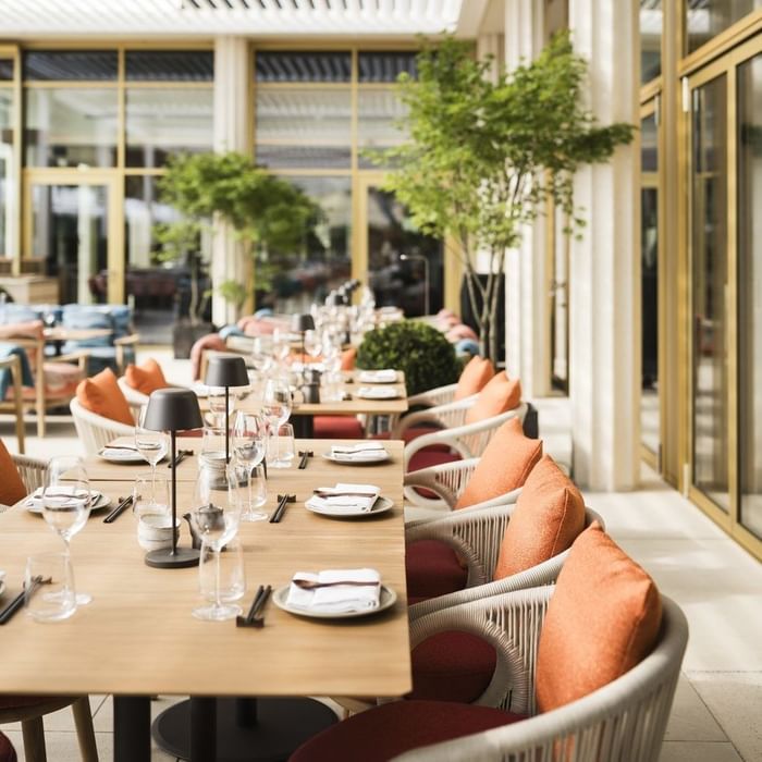 Elegantes Restaurant mit gedeckten Tischen, weißen Stühlen, orangen Kissen und grünen Pflanzen.