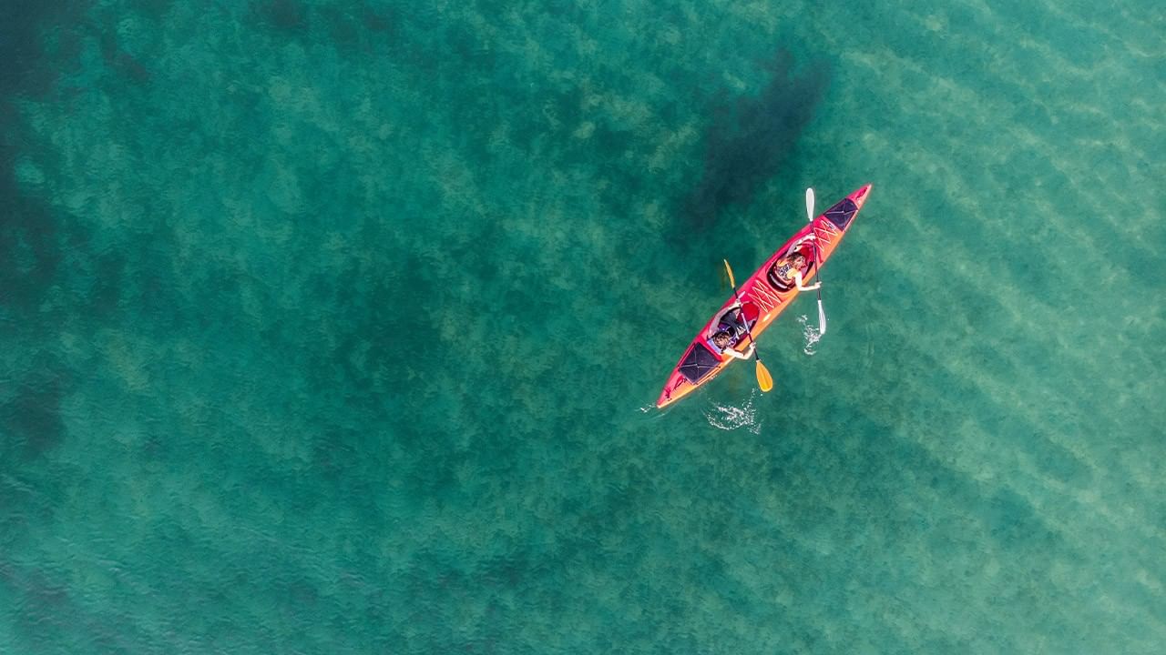 Two people in a red kayak with paddles on green water from above.