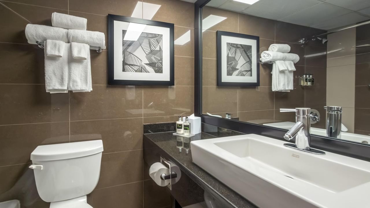 Modern bathroom with brown tile walls, white fixtures, black countertop, and framed artwork at Coast Calgary University Area Hotel by APA