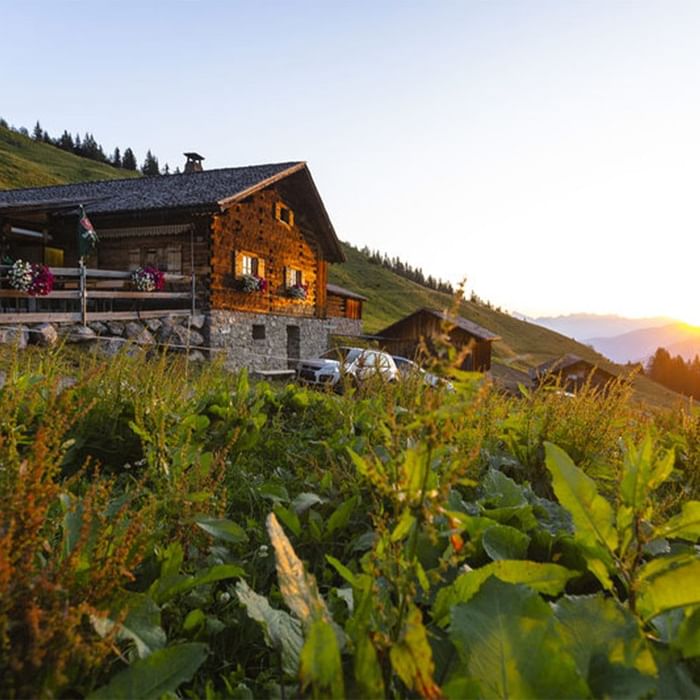 Sonnenuntergang über einem traditionellen Holzhaus in einer grünen Berglandschaft mit Autos und Pflanzen.