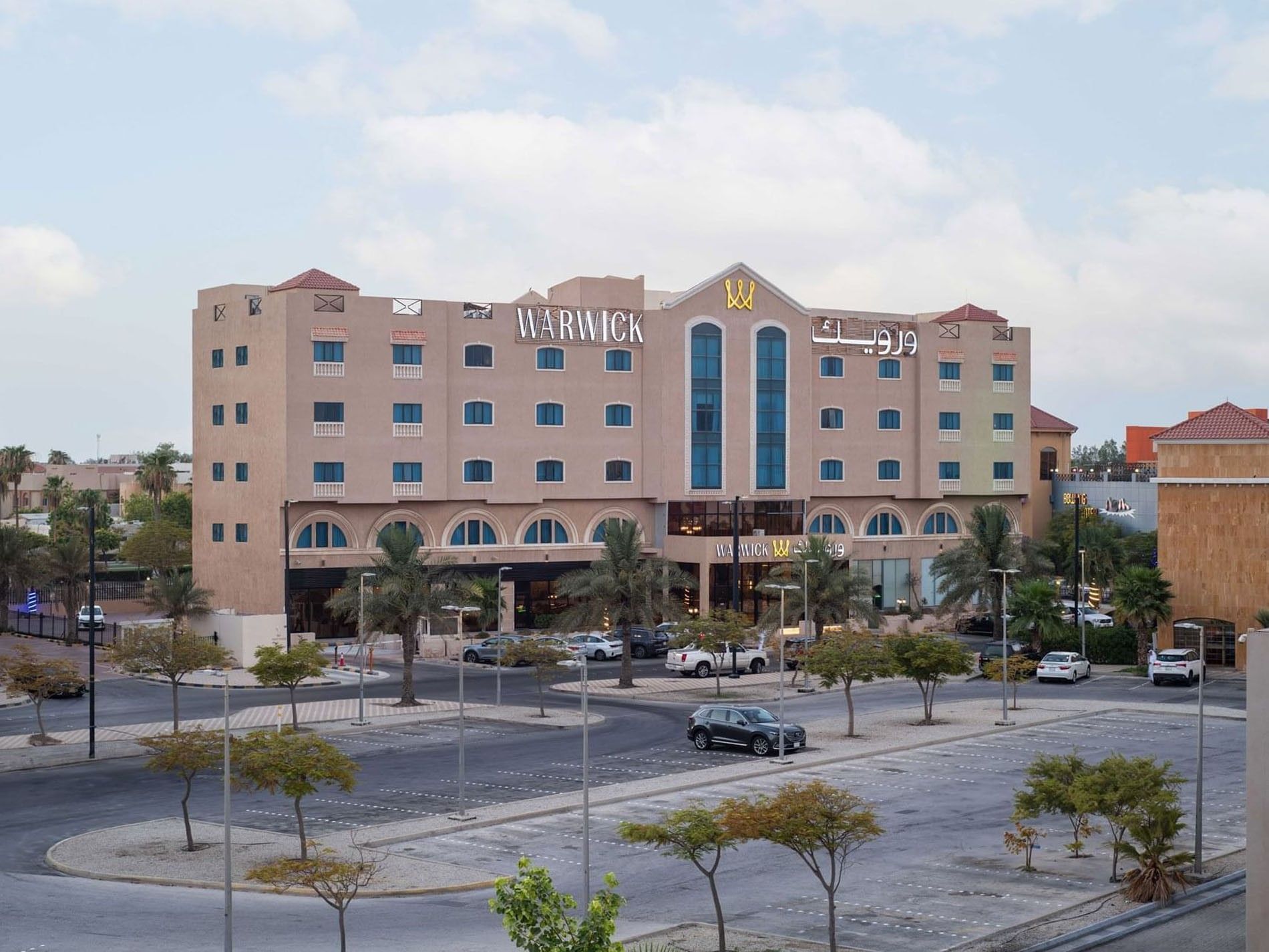 Multi-story facade of Warwick Al Jubail by a paved parking lot surrounding green trees and street lamps in a city