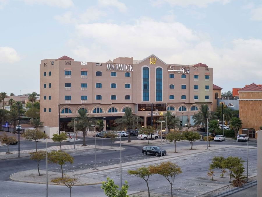 Multi-story facade of Warwick Al Jubail by a paved parking lot surrounding green trees and street lamps in a city