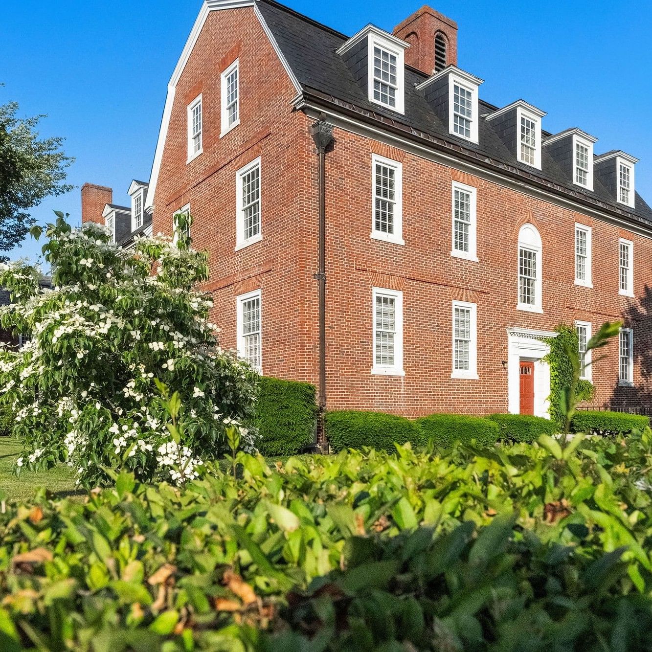 Red brick colonial house with multiple windows, a flag, and lush greenery under a clear blue sky.