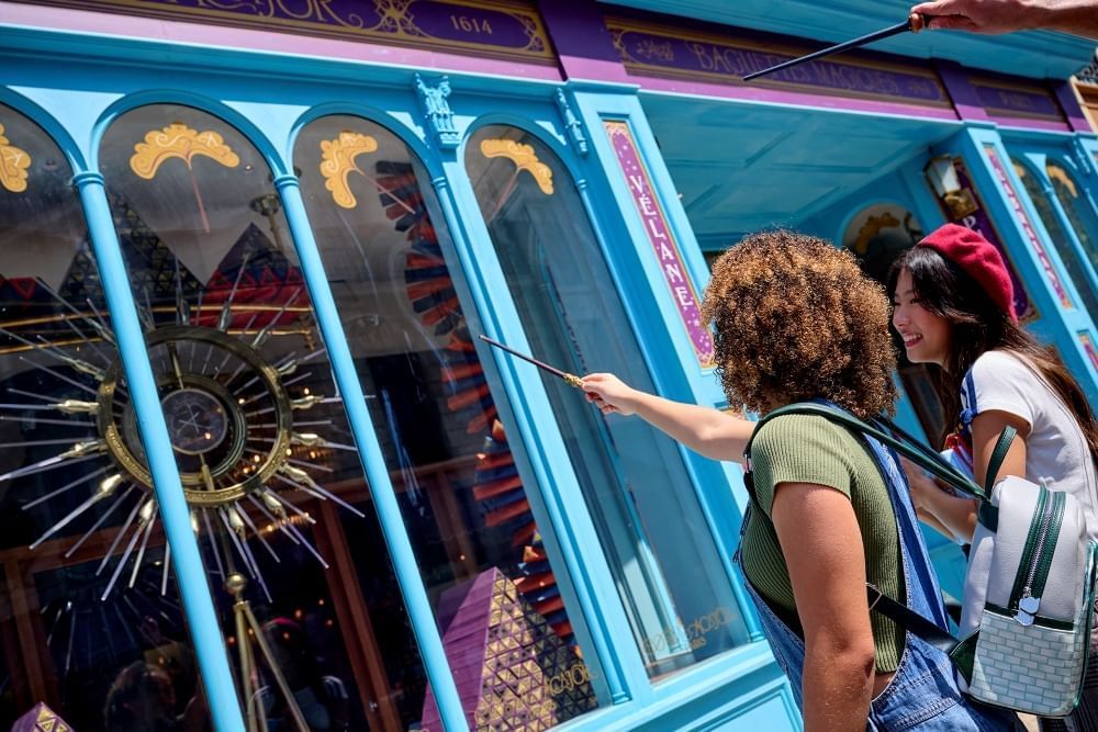 Two women, one pointing a wand, stand in front of a store front with arched windows and light blue walls. 