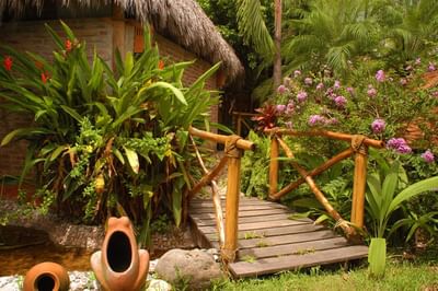Wooden bridge over small pond with plants, flowers, and clay sculptures in a lush garden setting.