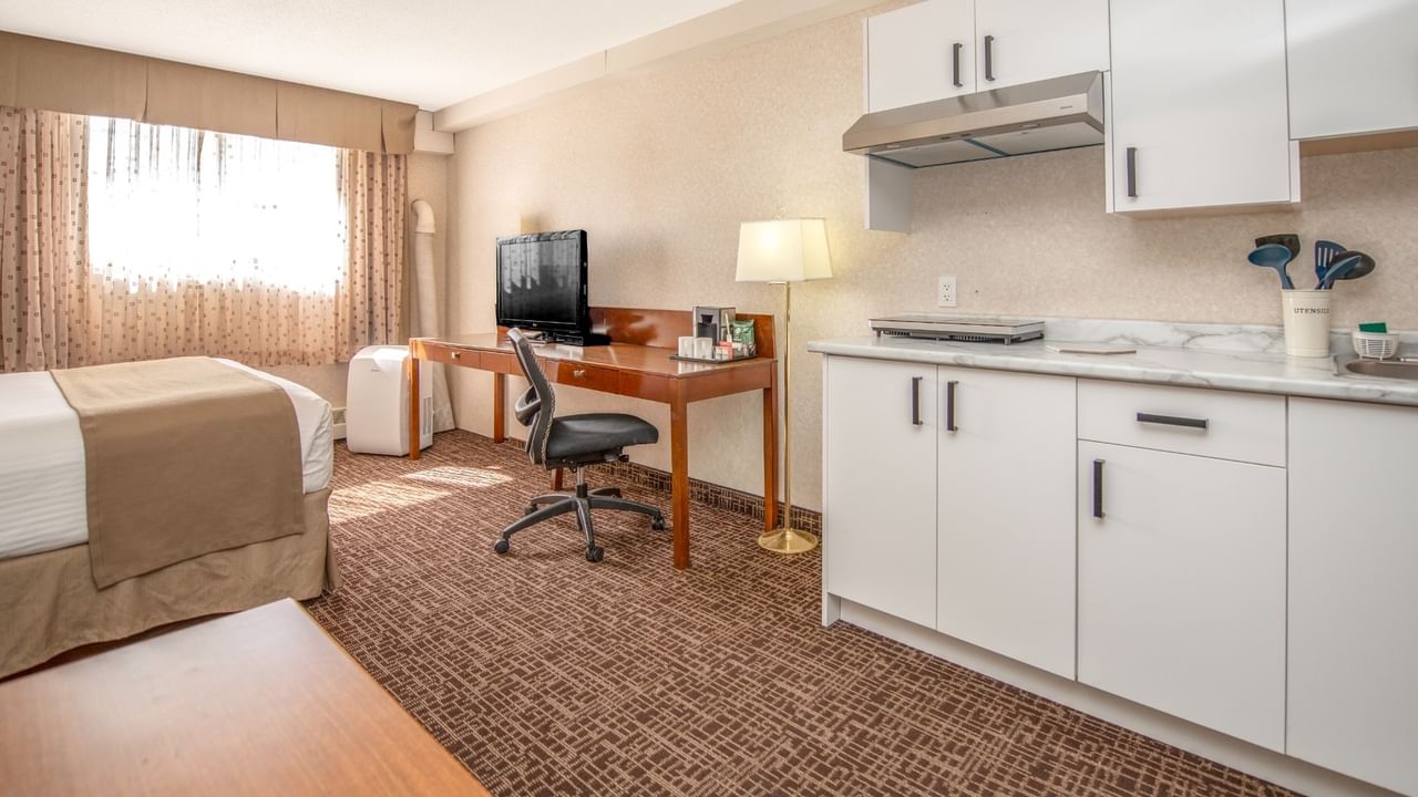 A hotel room with a bed, desk, chair, TV, and a small kitchen area.