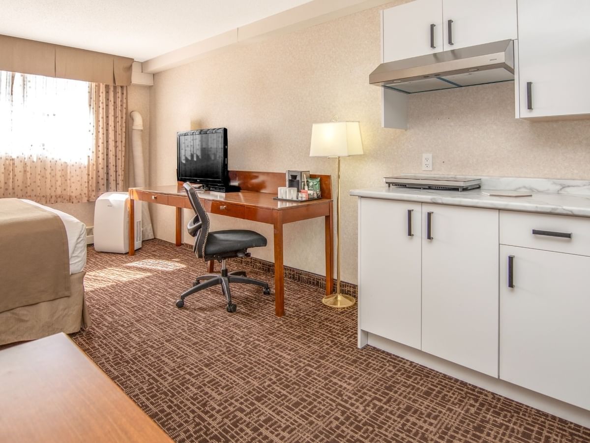 Hotel room with bed, desk, TV, chair, lamp, and kitchenette with cabinets and countertop.