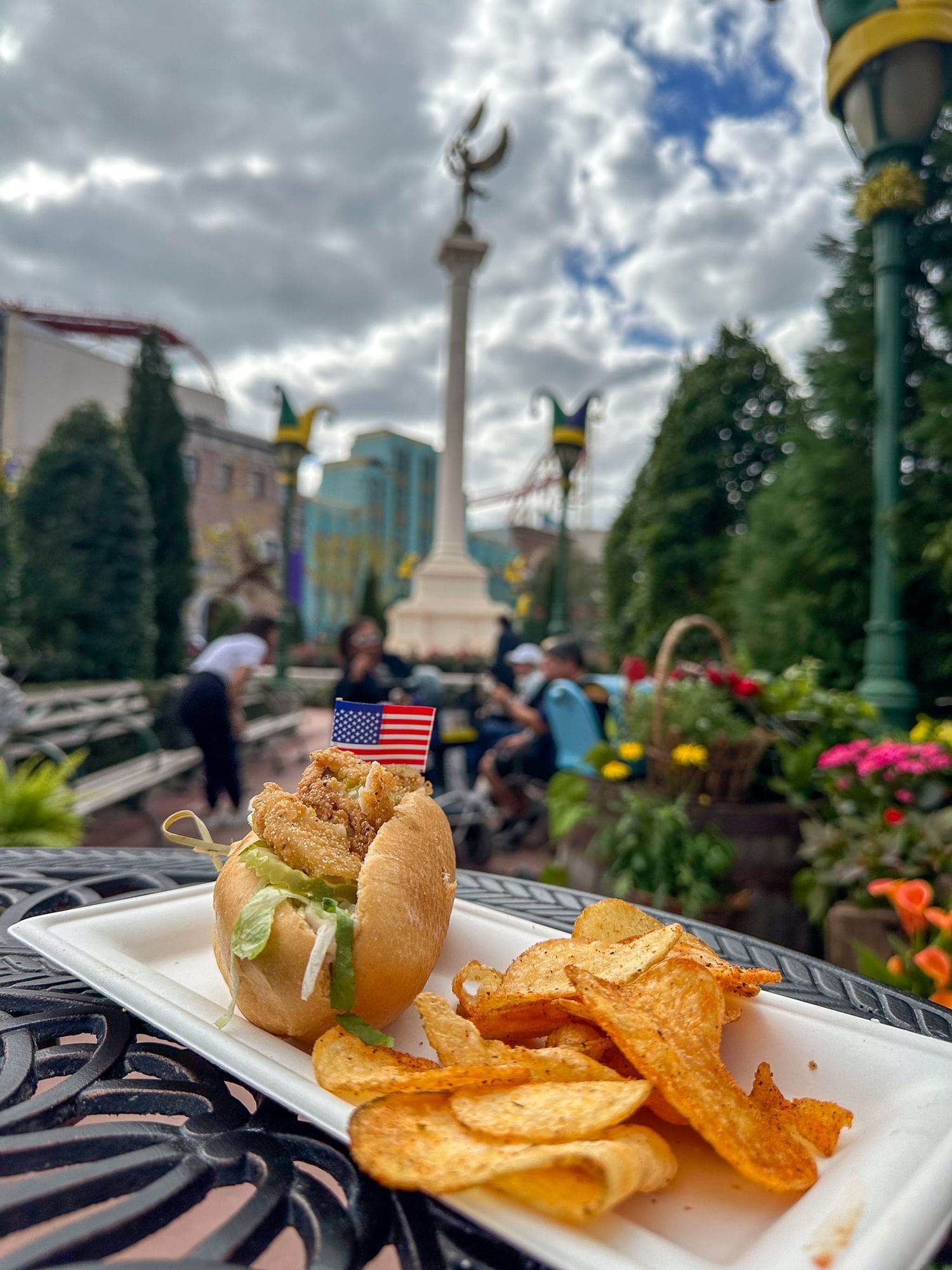 A fried shrimp po'boy sandwich with a tiny American flag and potato chips on a white plate, set against a backdrop of Universal Orlando's Mardi Gras celebration with trees, flowers, and people.