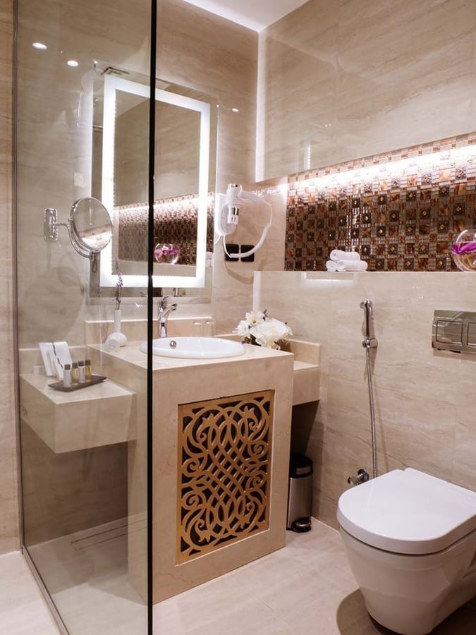 Beige bathroom with a sink by a toilet under a lit mirror in Deluxe Room at Cantonal Hotel by Warwick Riyadh