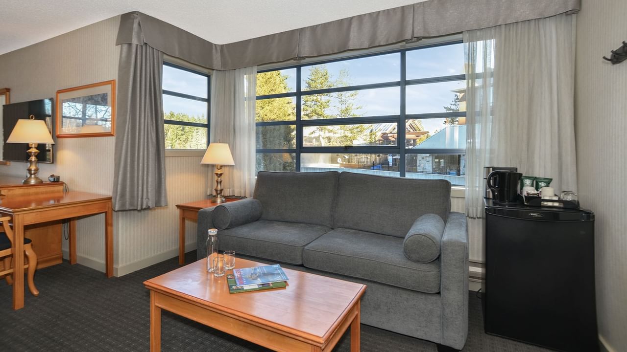 Spacious hotel suite with sofa, coffee table, mini fridge, and large windows.