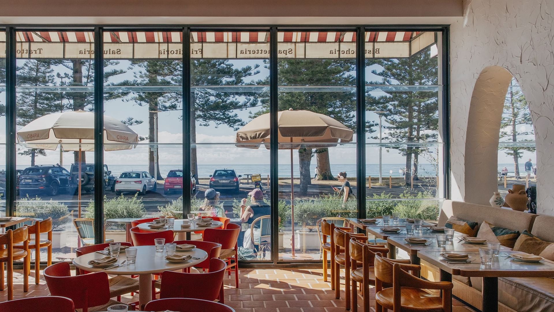 Chairs set by tables in a restaurant, looking through windows under striped awnings toward the sea at Elysium Manly