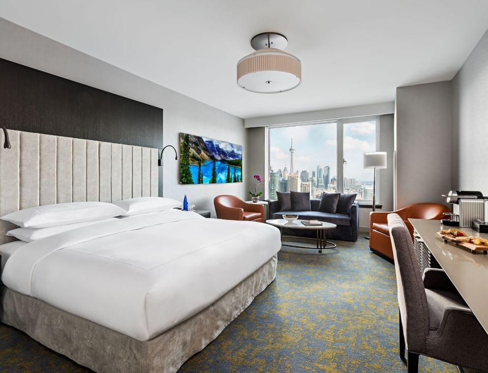 King bed set by a mountain artwork, leather chairs and sofa near windows overlooking the city skyline at Hotel X Toronto