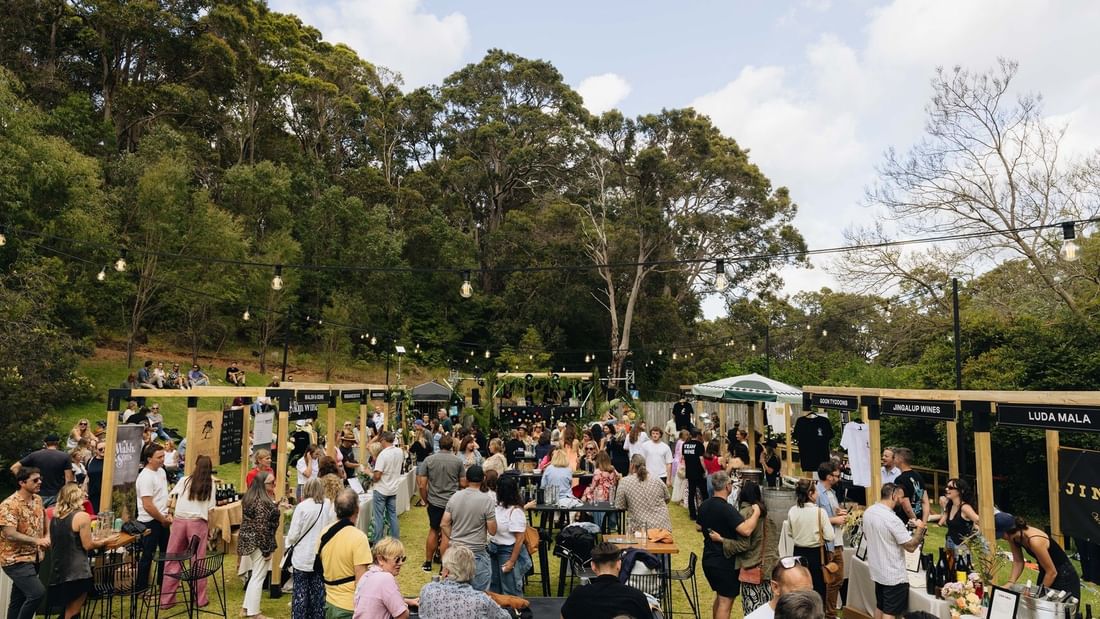 Outdoor wine and food event with attendees mingling among vendor booths under string lights and trees.