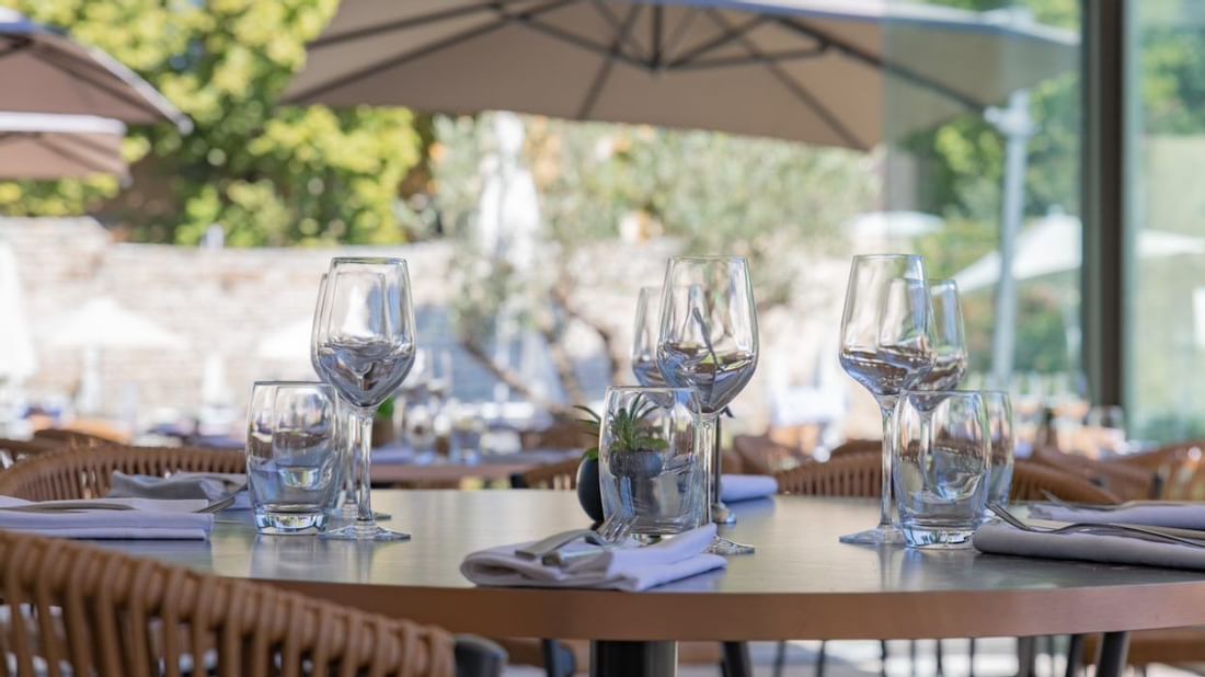 Table de restaurant avec verres et couverts, sous une terrasse ombragée avec chaises et parasols.