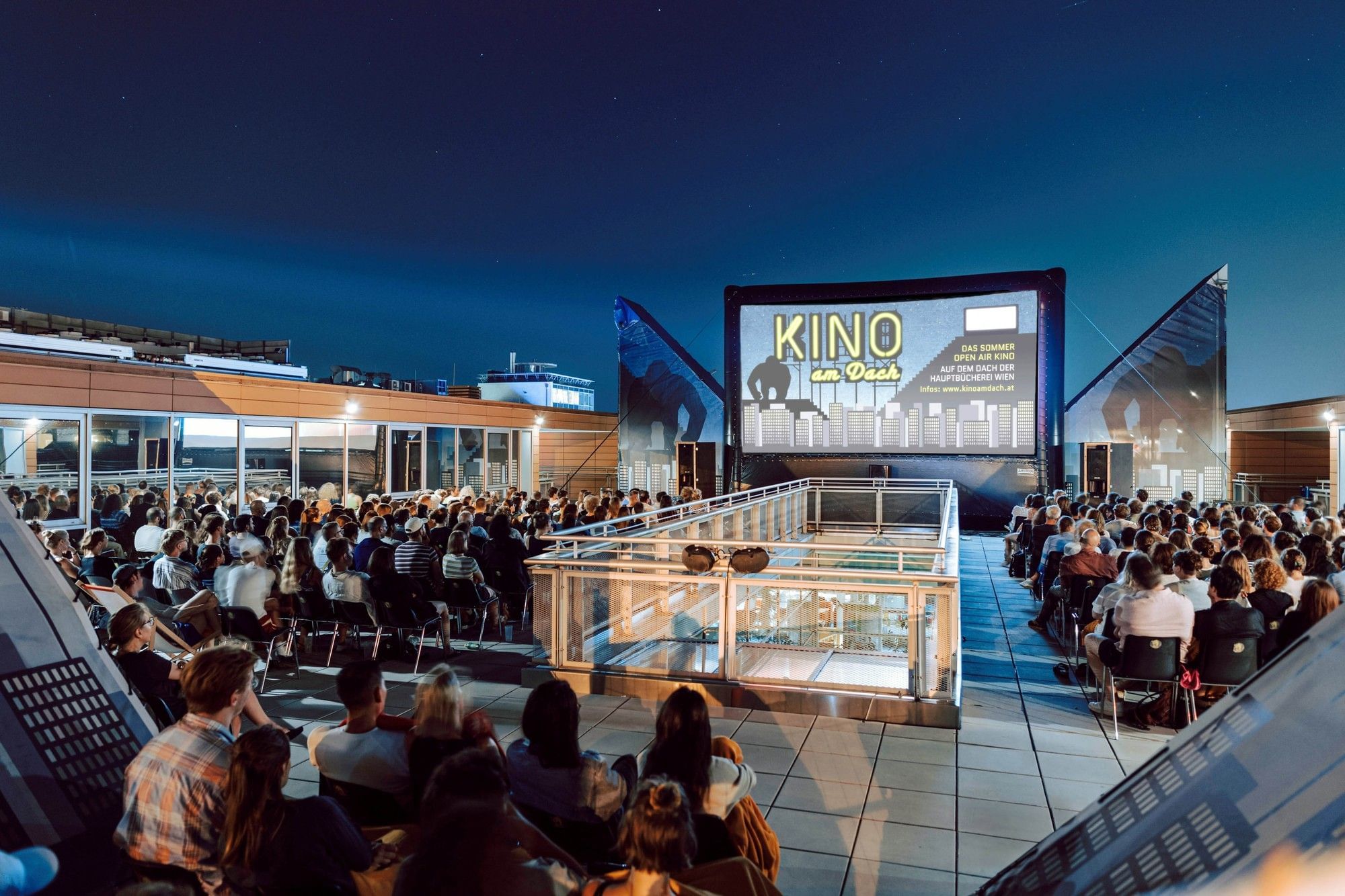 Besucher genießen Kino am Dach in Wien unter freiem Himmel mit Blick auf die Stadt und großer Leinwand bei Abendstimmung.