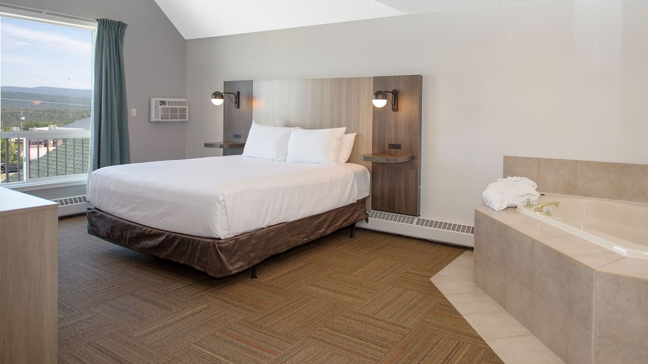 Hotel room with bed, bathtub, window, and balcony overlooking mountains and buildings.