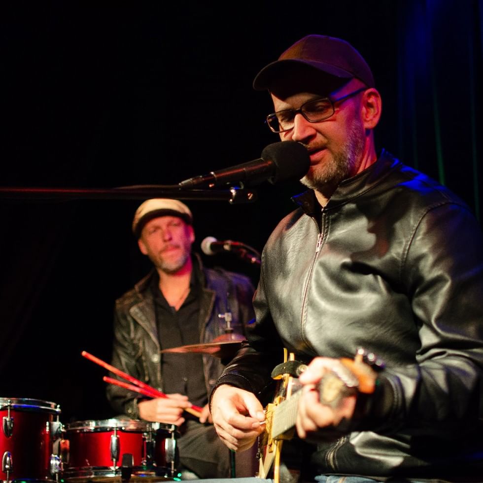 Two men in black leather jackets, one playing guitar, the other playing drums, both on stage.