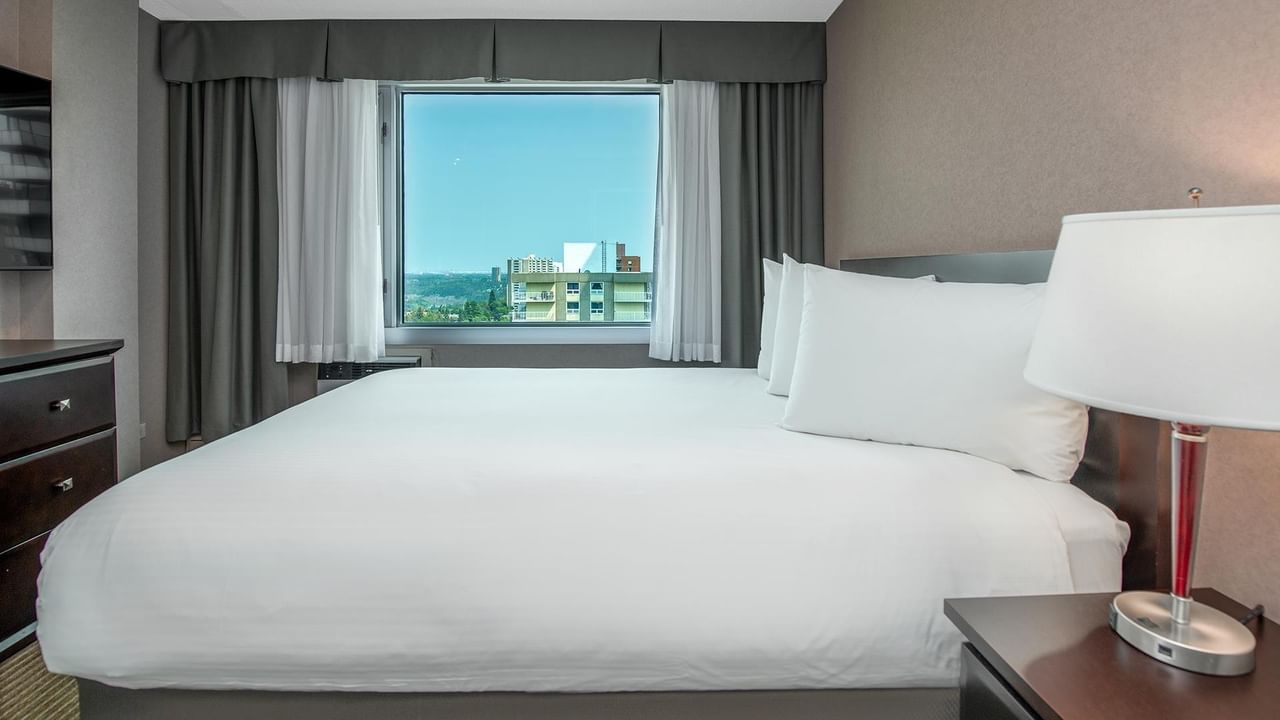 Hotel guest suite with a queen bed, bedside lamp and large window with a city view.