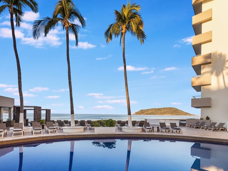 Sunvivia Mazatlán resort pool view facing the ocean with palm trees, lounge chairs, and a distant island hill