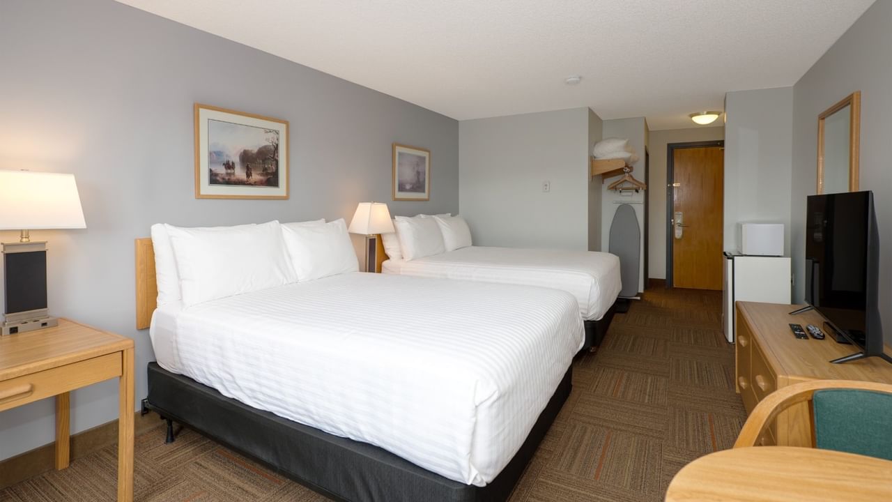 Two beds, lamps, table, TV, and door in a hotel room with gray walls.