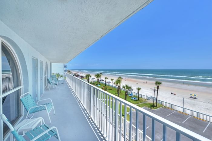 A balcony with a white railing and blue chairs overlooking a beach with palm trees and a parking lot.