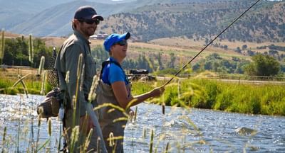 Two people fish by a river surrounded by tall grass and mountains near Hotel Park City Autograph Collection