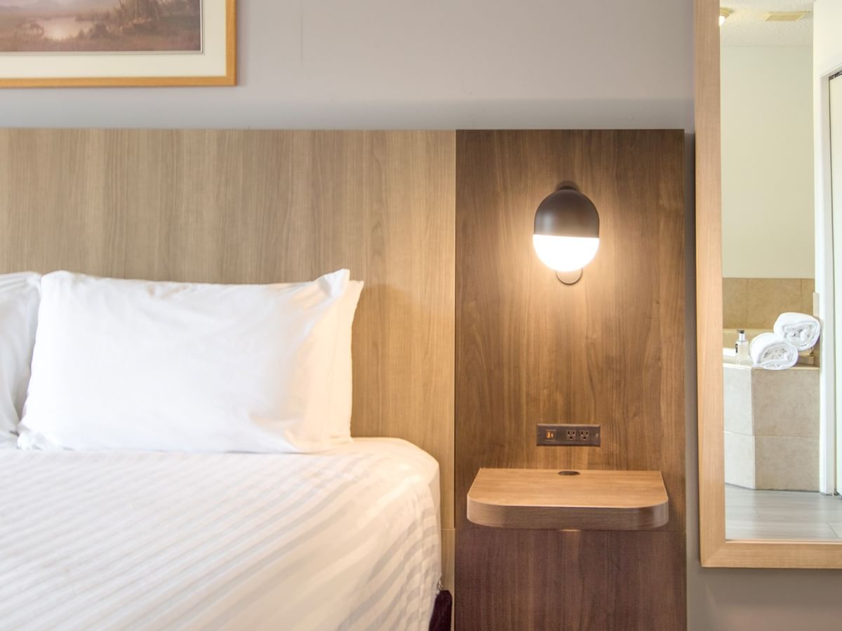 A hotel room with a bed, wooden headboard, lamp, and mirror reflecting a bathroom.
