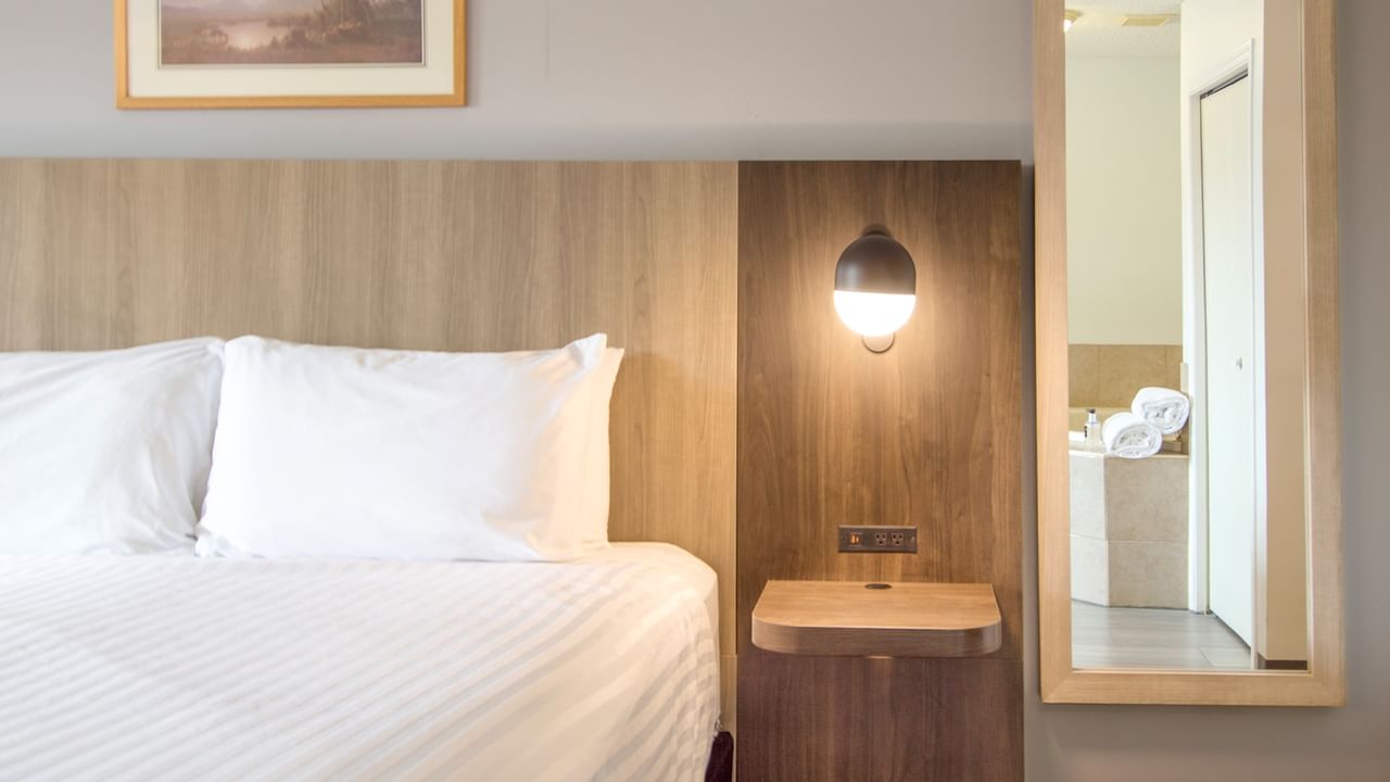 A hotel room with a bed, wooden headboard, lamp, and mirror reflecting a bathroom.