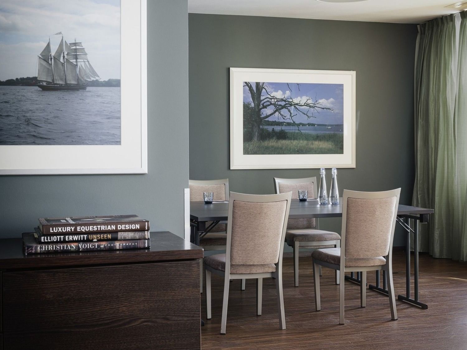Esszimmer mit dunklen Holzböden, grauen Wänden, einem Holztisch und gerahmten Gemälden von Meereslandschaften und Bäumen.