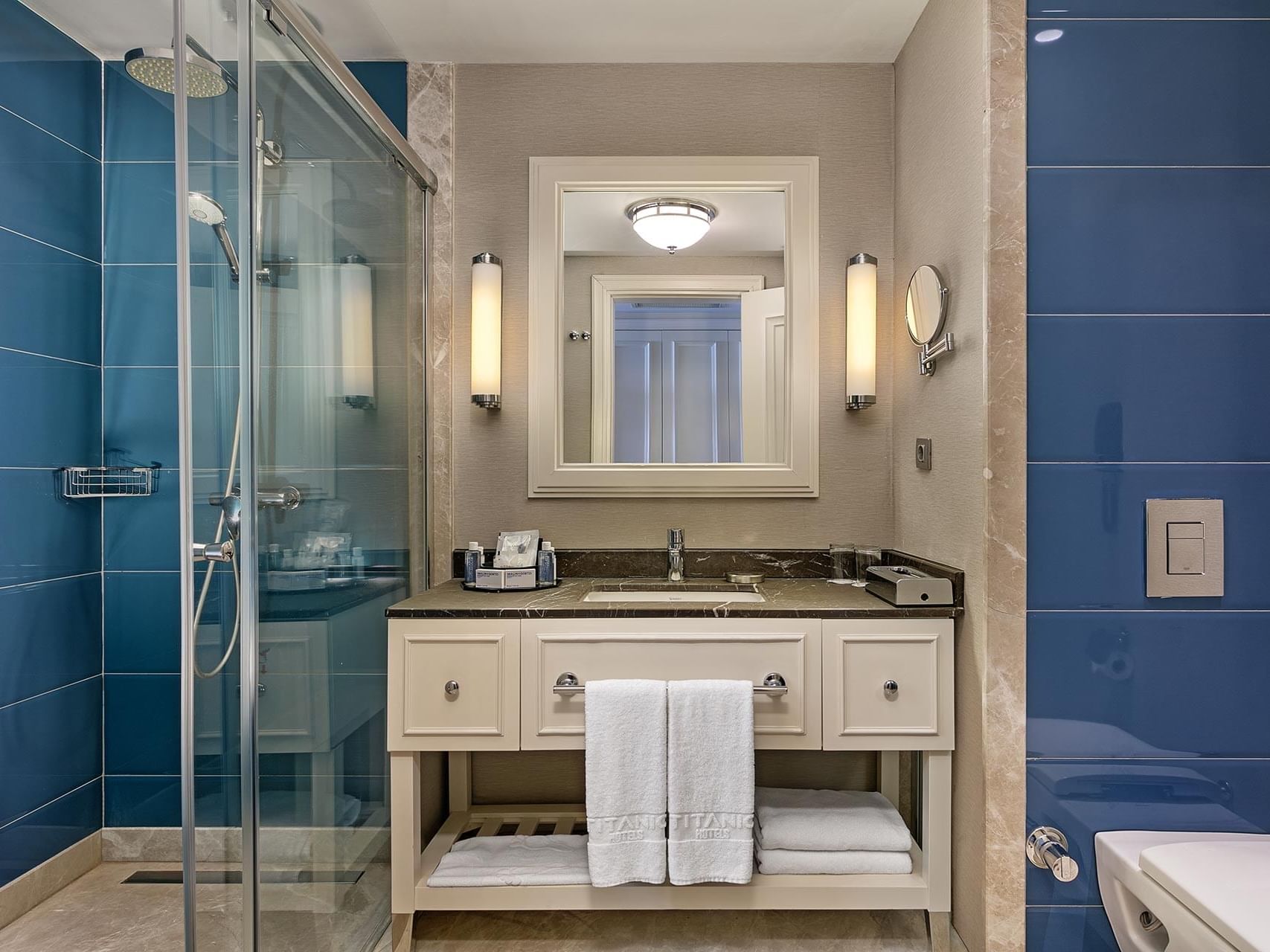 Modern bathroom with blue tile, glass shower, white vanity with dark countertop, and elegant wall sconces at Titanic Deluxe Golf Belek