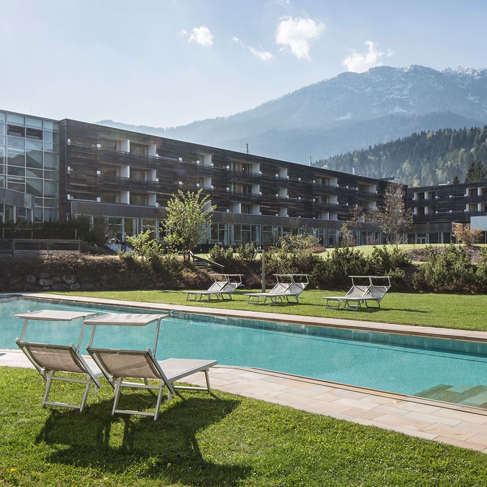 Piscina all'aperto con lettini, vicino a un edificio moderno e montagne sullo sfondo.