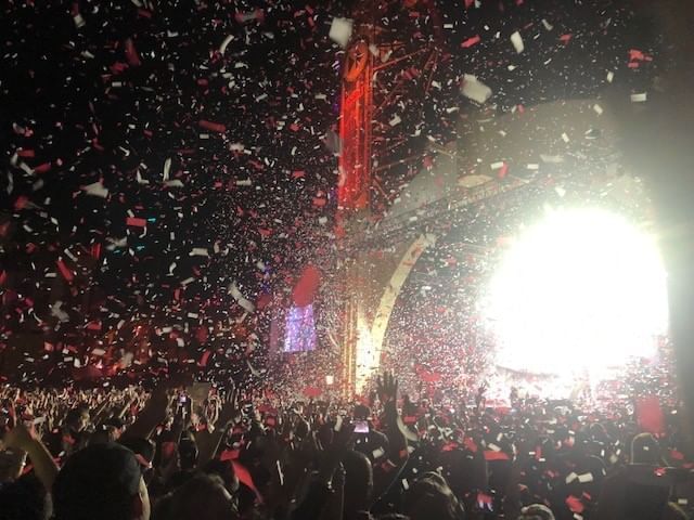 A bright light on a stage glows at night over a large crowd and a sky of falling confetti. 