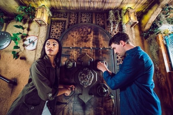 A man and woman stand in front of a large wooden door. The man is attempting to unlock a padlock while the woman looks out with a look of surprise. 