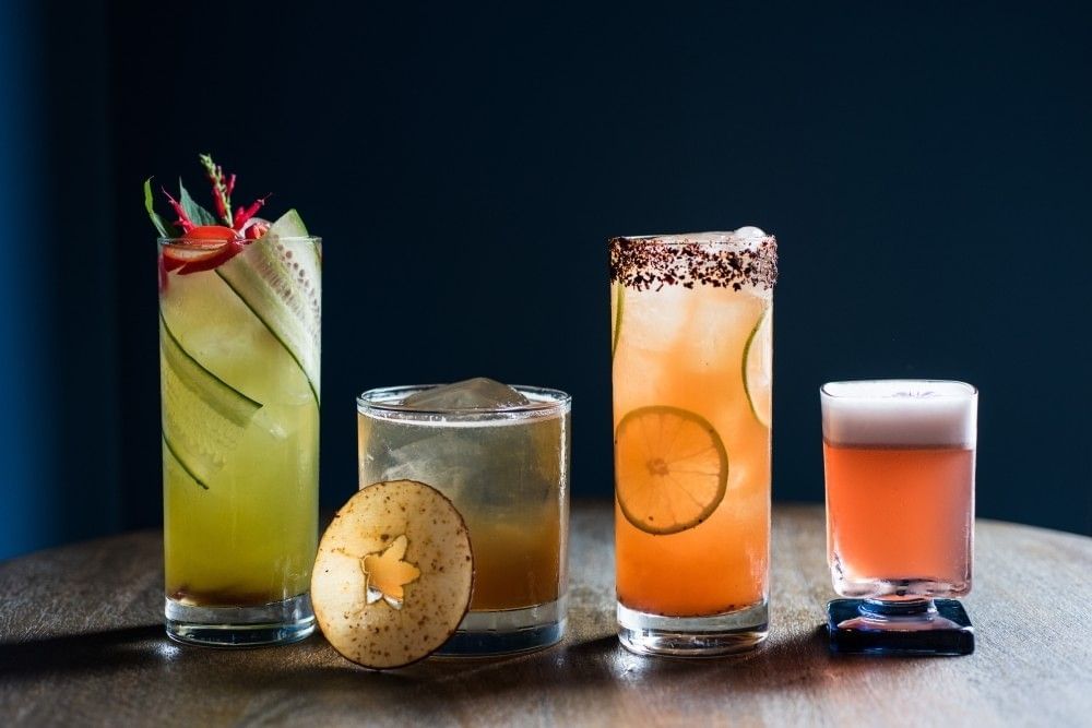 Four colorful and creative cocktails sit on a sleek wooden surface against a dark backdrop.