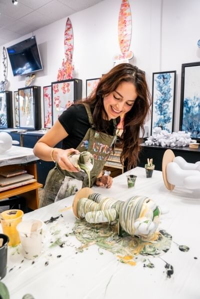 A woman in an apron pours marbled paint over a figurine in a studio decorated with abstract artwork.