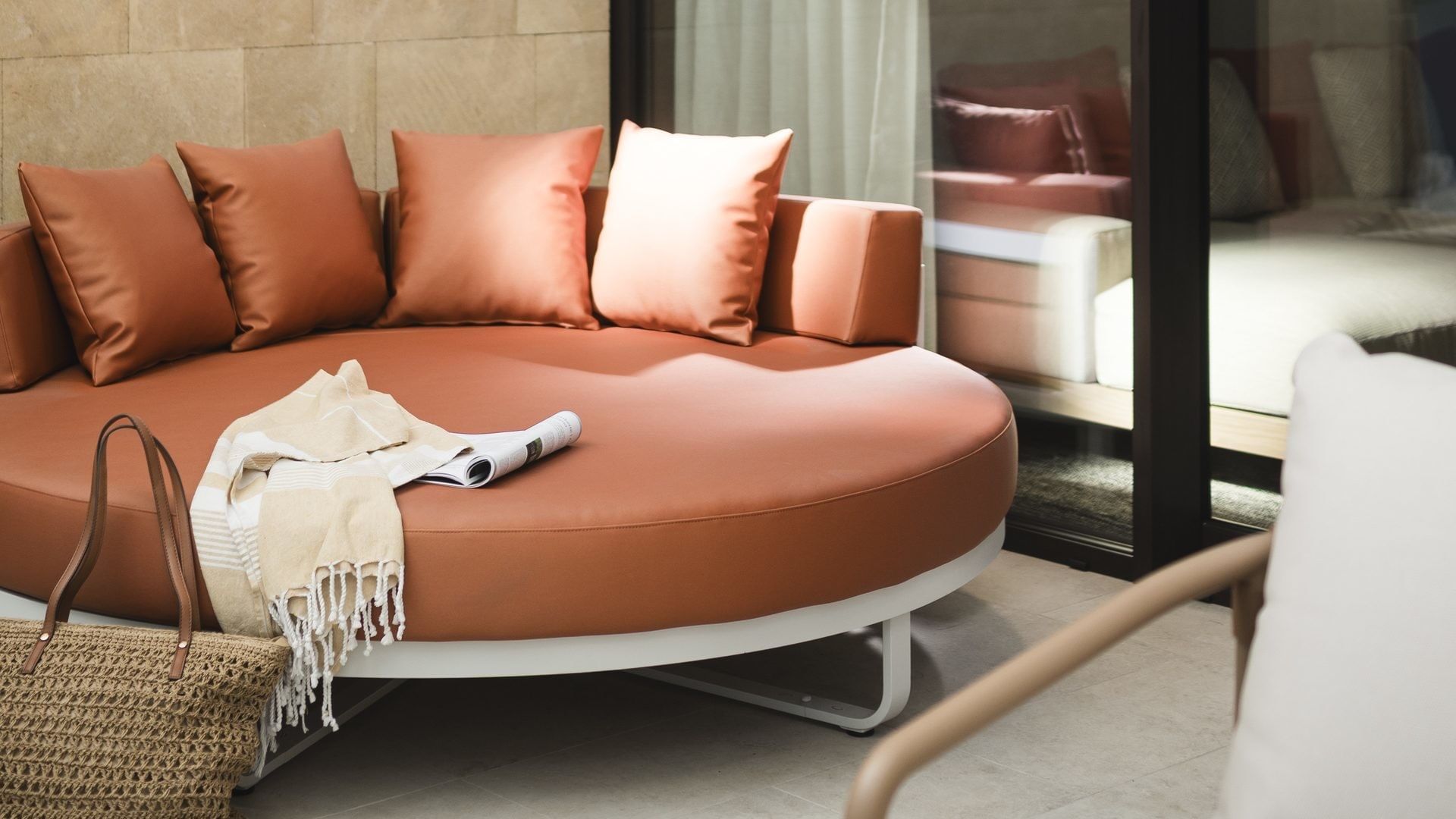 Divano rotondo marrone con cuscini, borsa, sedia e riflessione di un letto in vetro.