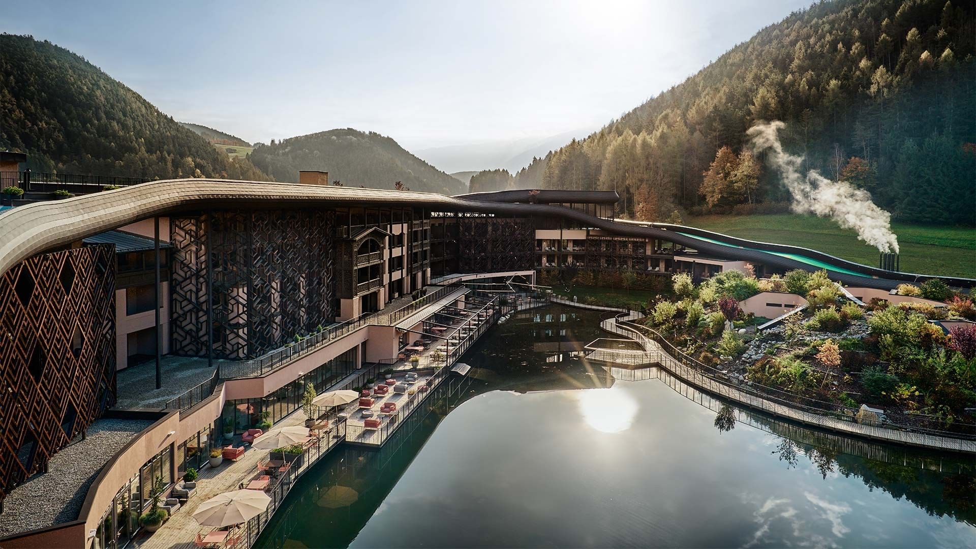 Aerial view of a modern hotel with a pool, surrounded by mountains and greenery.