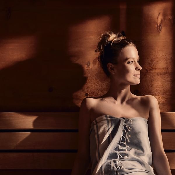 Frau in Badetuch auf einer hölzernen Saunabank, blickt nach oben, Sonnenlicht erzeugt Schatten.