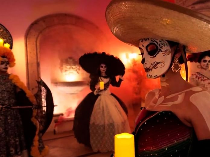 Performers in traditional Day of the Dead face paint and costumes gather in a warmly lit room with candles and decorative arches.