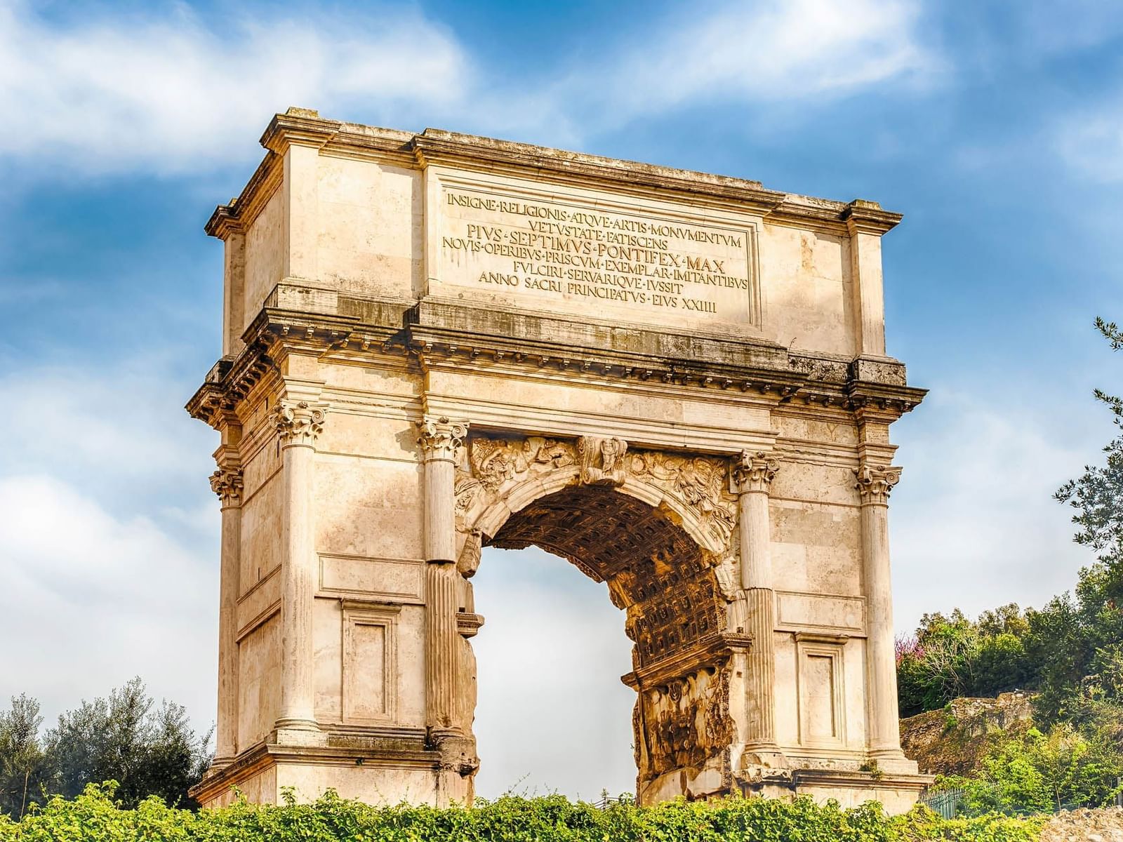 The Arch of Titus with intricate carvings, set against a bright sky and greenery near The Glam Hotel