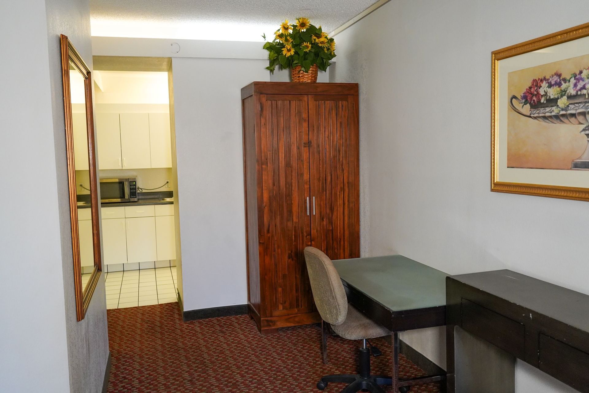 A hotel room with a desk, chair, cabinet, and kitchenette.