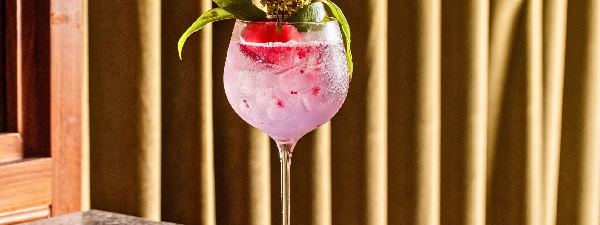 Glass of pink cocktail with fruit and leaves garnish, on a table with a curtain in the background.