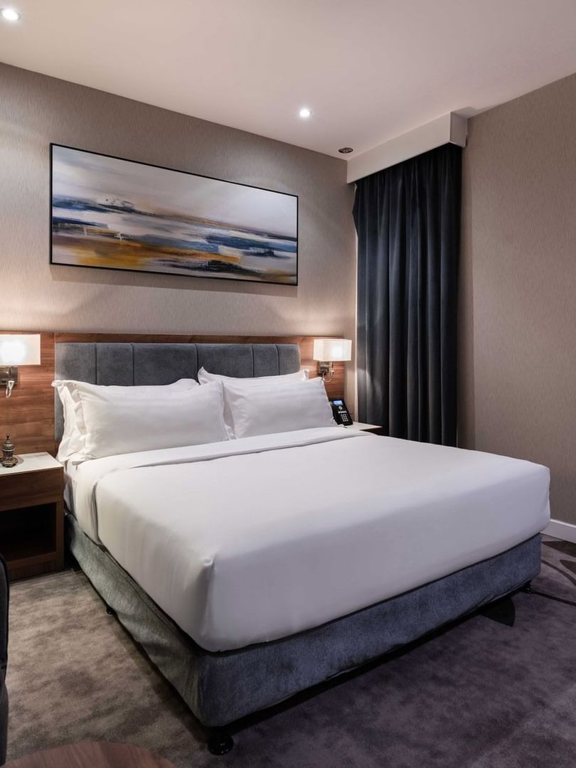 Plush bed by a grey armchair and tall mirror under a landscape painting in Standard Room at Warwick Al Jubail