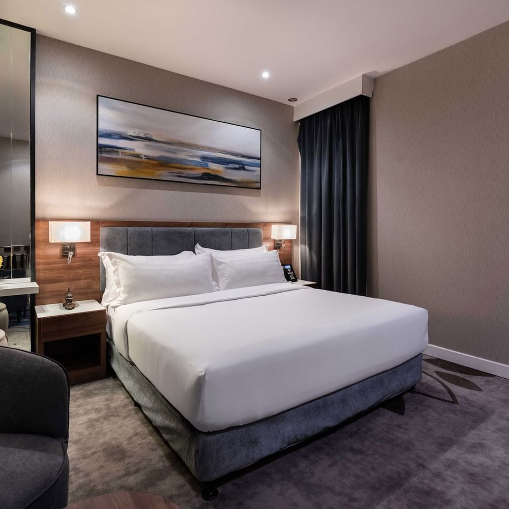 Plush bed by a grey armchair and tall mirror under a landscape painting in Standard Room at Warwick Al Jubail