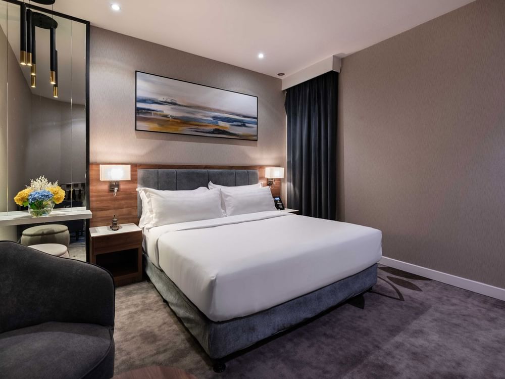 Plush bed by a grey armchair and tall mirror under a landscape painting in Standard Room at Warwick Al Jubail