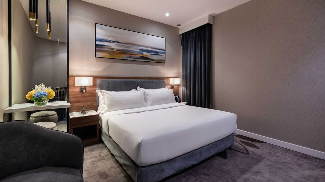 Plush bed by a grey armchair and tall mirror under a landscape painting in Standard Room at Warwick Al Jubail