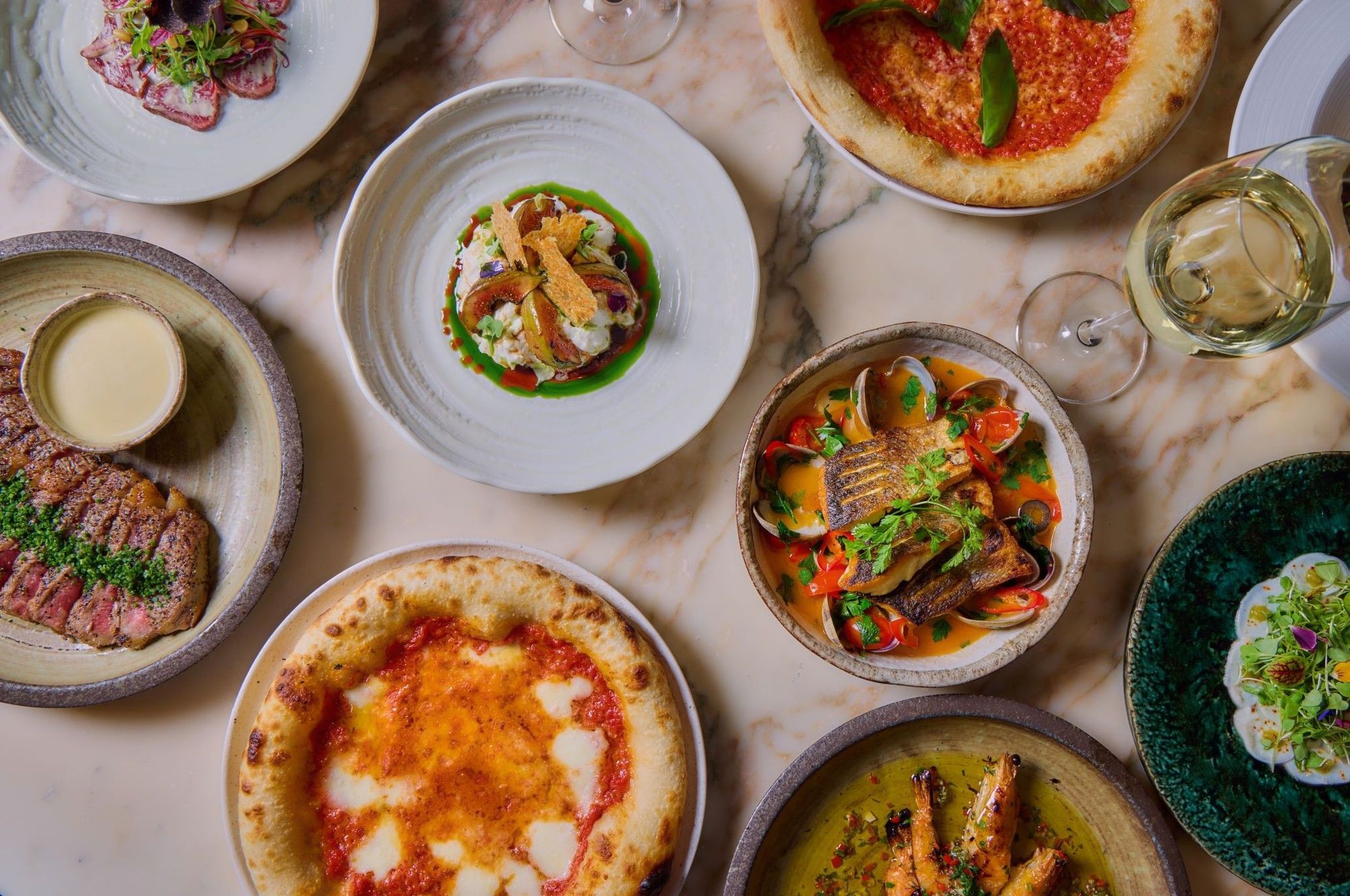 Assorted gourmet dishes from the A La Carte menu and two wine glasses on a marble table in the May Fair Kitchen