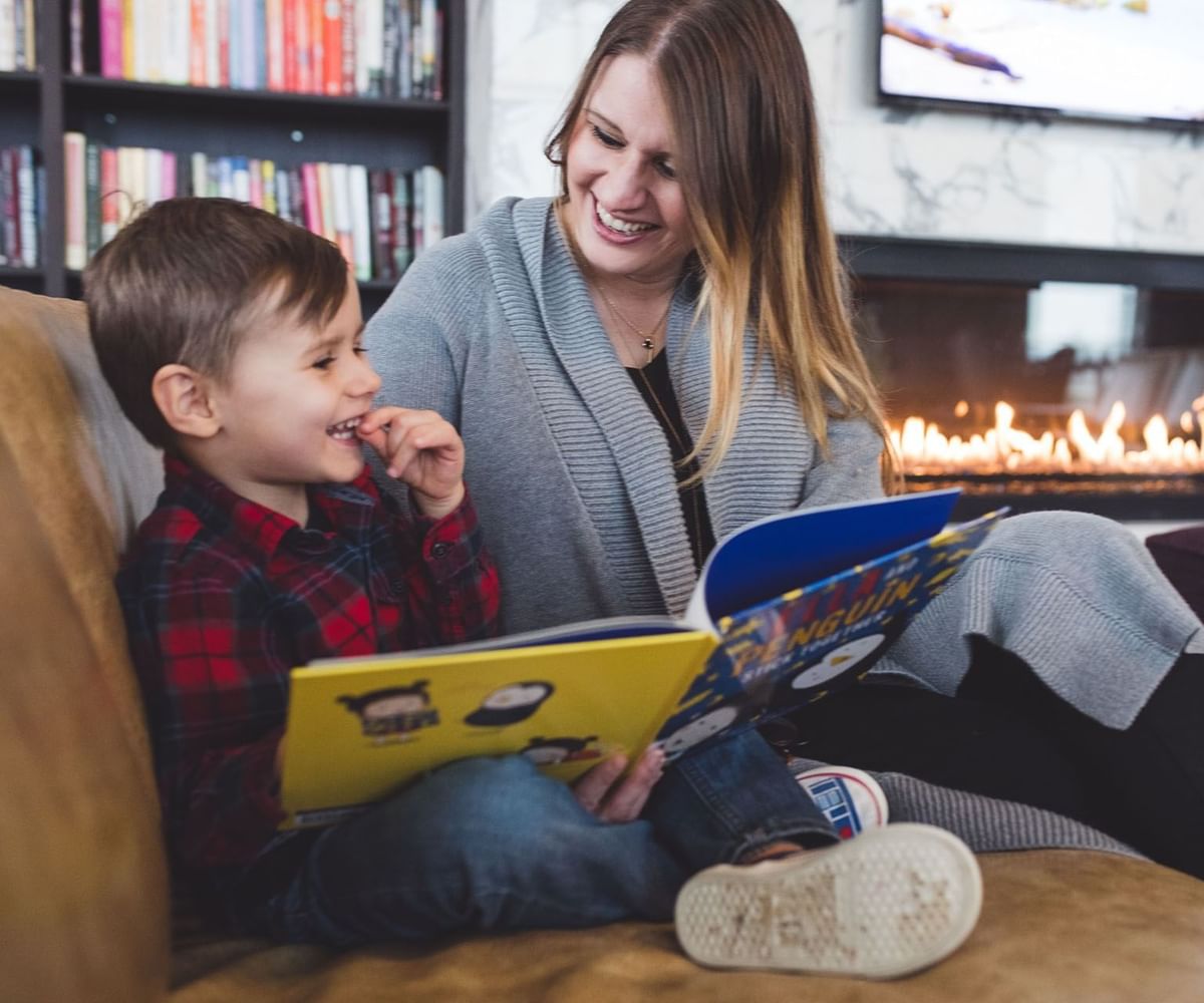Mother and young child reading colorful book together on sofa facing warm fireplace at Hotel X Toronto