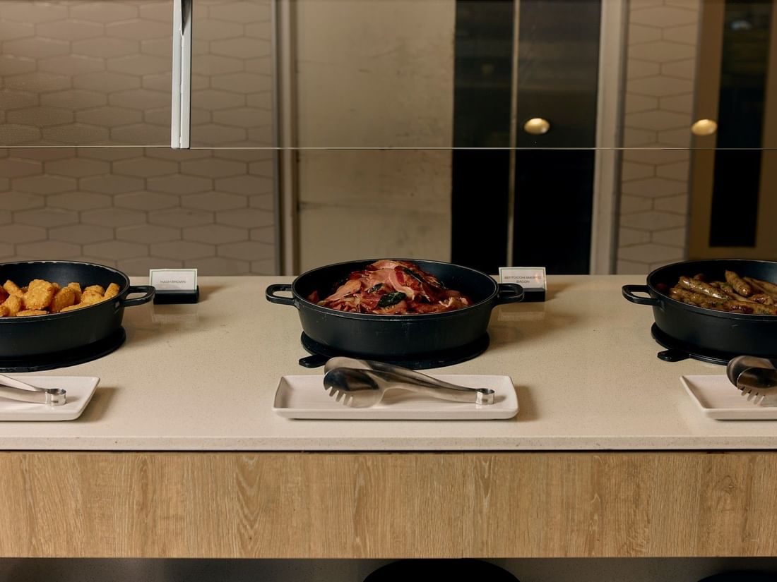 Hot breakfast items including hash browns, bacon, and sausages in black skillets at Pullman East Melbourne