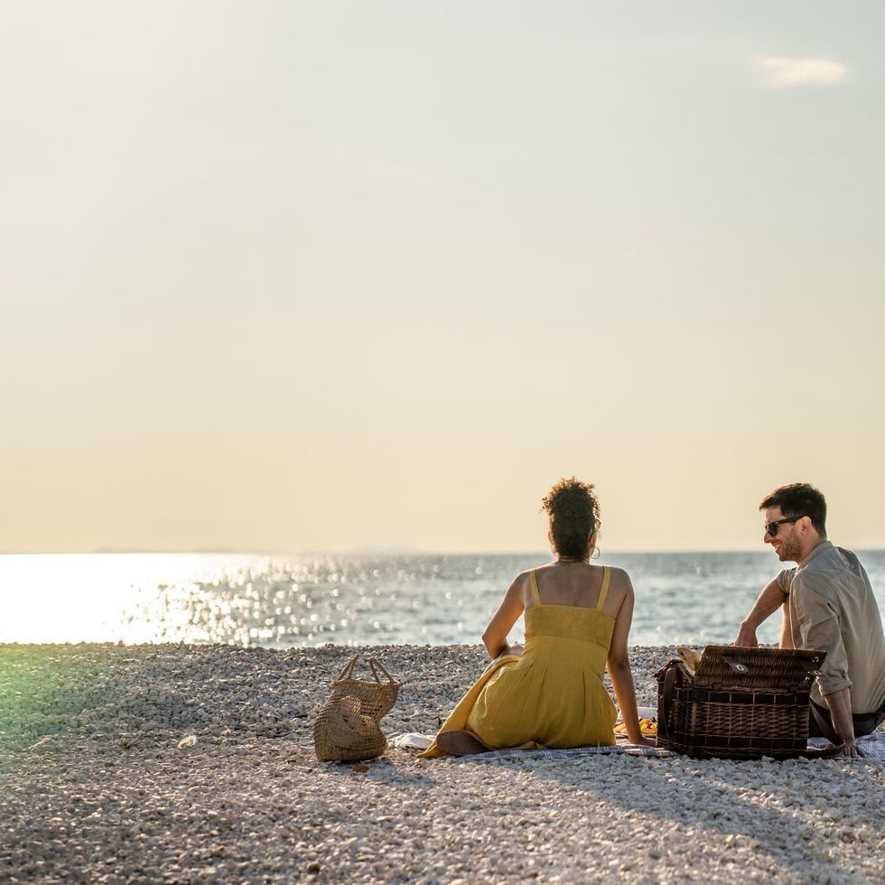 Zwei Personen sitzen am Strand mit Körben und Meerblick bei Sonnenuntergang.